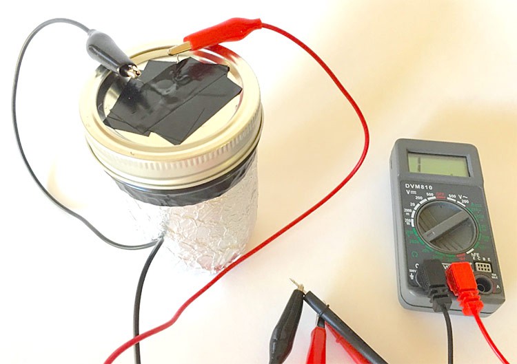 A multimeter connects to the lead wires of a photoresistor that is placed in the lid of a mason jar