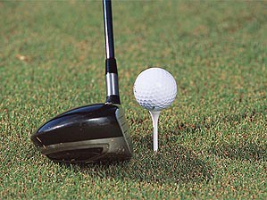 The head of a driver rests below a golf ball on a tee