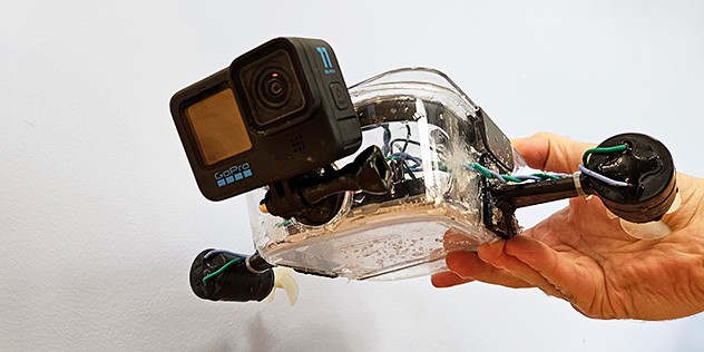 A clear plastic container with electronics inside, two motors mounted so the sides, and a GoPro mounted to the front