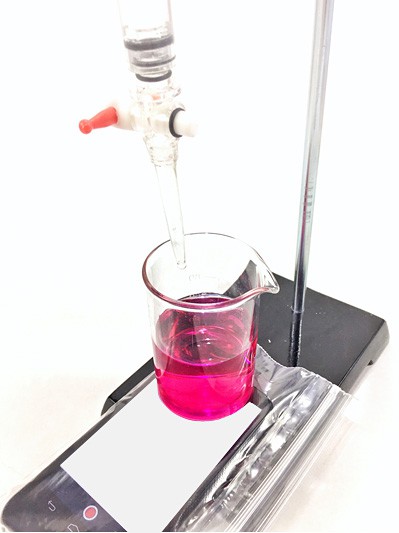 A smartphone in a plastic bag rests under a beaker filled with pink liquid