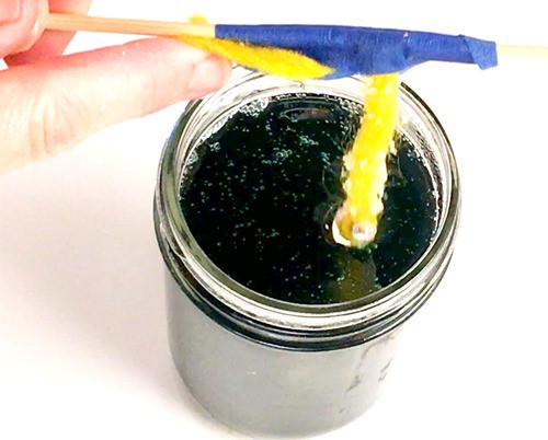 Person holding a skewer with a sugar-coated string attached. The string hangs down into a jar which contains a thick solution. 