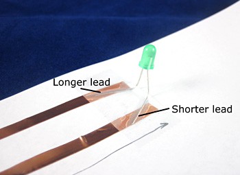Leads of an LED are bent and taped to different strips of copper tape on a piece of paper