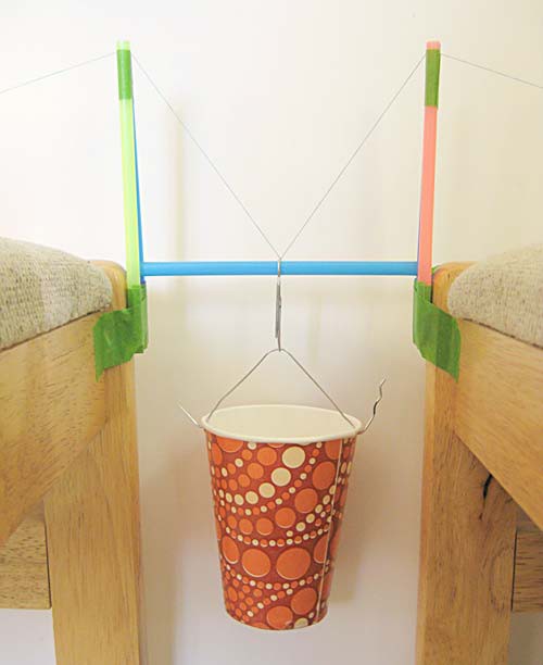 A paper cup is suspended from the center of a straw that is supported by two tensioned threads from a tower bridge