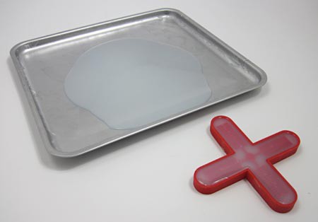 A red cross-shaped mold filled with silicon next to a puddle of silicon on a baking sheet
