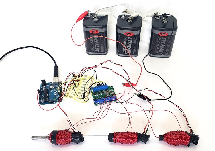 An Arduino, a circuit board, three lantern batteries, and a clear plastic tube with three coils of wire around it. 