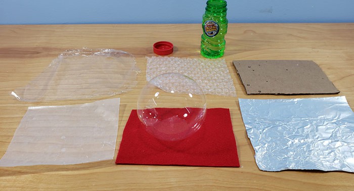 A bubble rests on a square of felt next to squares of plastic wrap, bubble wrap, cardboard, wax paper and aluminum foil