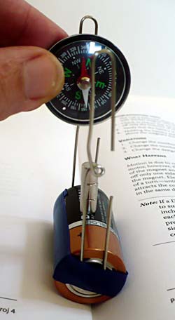 A compass is held above a stack of three magnets between two paperclips that are attached to the terminals of a battery