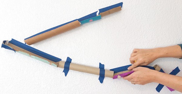 Several cardboard tubes taped to a wall to make a marble run. A person is rearranging one cardboard roll with her hands.