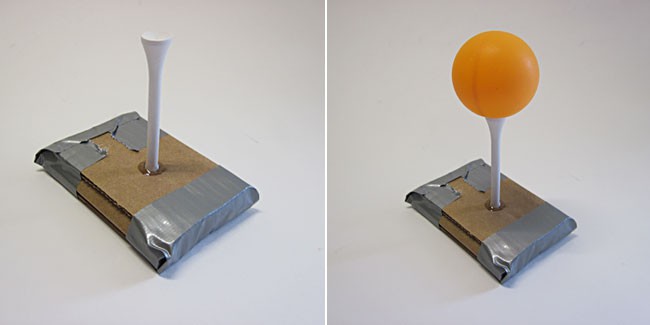 A golf tee stuck into cardboard holds up a ping pong ball