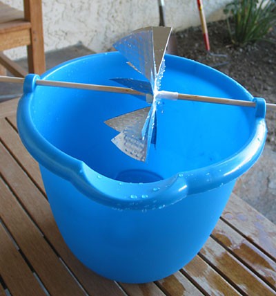 A waterwheel cut from an aluminum pie pan held over a plastic bucket by a wooden dowel
