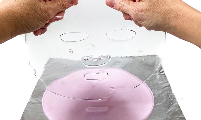 Two hands hodling a transparent hydrogel face mask above a face mask mold. 