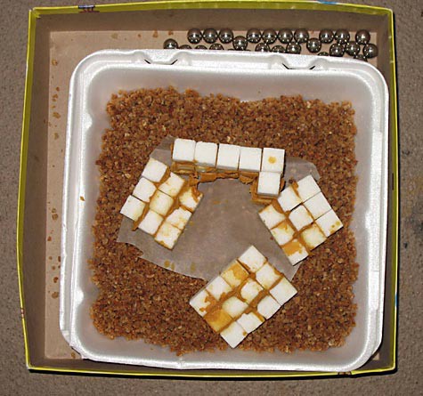 A collapsed structure made of sugar cubes and peanut butter sits in a tray of coarsely ground cereal