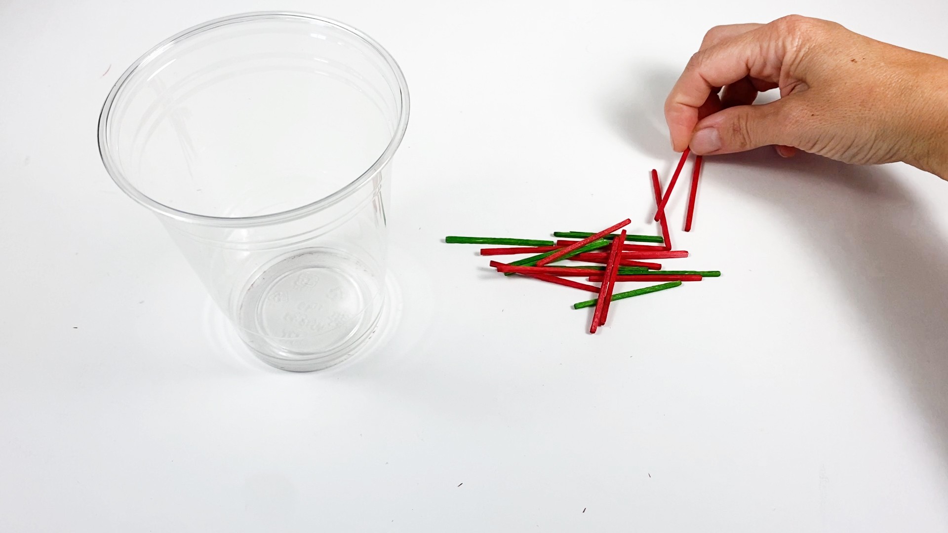 A pile of green and red matchsticks next to a plastic cup. A hand is holding one of the red matchsticks.